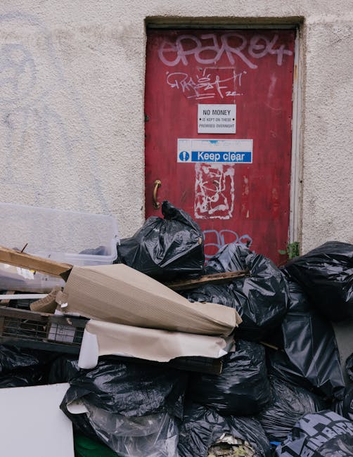 The image shows a cluttered outdoor area with a red door covered in graffiti, set within a textured, off-white concrete wall. In front of the door, there is an accumulation of black garbage bags, some torn open revealing waste materials, and discarded cardboard boxes. A white plastic container and a wooden pallet are also visible among the refuse. The scene indicates a state of neglect and the need for waste removal and cleaning. Cleaners Merton specializes in surface cleaning, deep cleaning, and sanitisation, providing professional services to maintain hygiene and cleanliness in both residential and commercial properties. Proper waste disposal and thorough cleaning of surfaces like floors, doors, and surrounding areas are essential for hygienic environments, especially after a clear-out or waste accumulation.