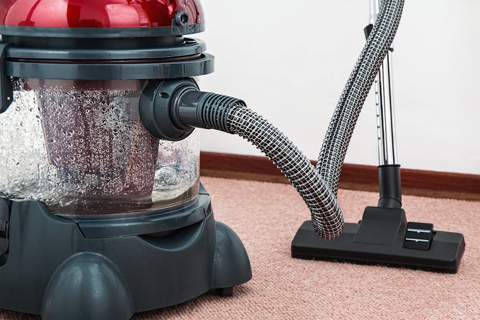 Close-up of a canister vacuum cleaner with a transparent dust container filled with debris, connected to flexible, patterned hoses, positioned on a pink carpeted floor within a room with plain white walls. The vacuum is turned on, with visible water droplets on the dust container indicating recent recent cleaning activity. The vacuum's base is black, and cleaning tools are attached nearby, suggesting deep surface cleaning and maintenance performed by Cleaners Merton in a domestic setting, aligned with surface sanitation and hygiene practices for residential carpet cleaning.