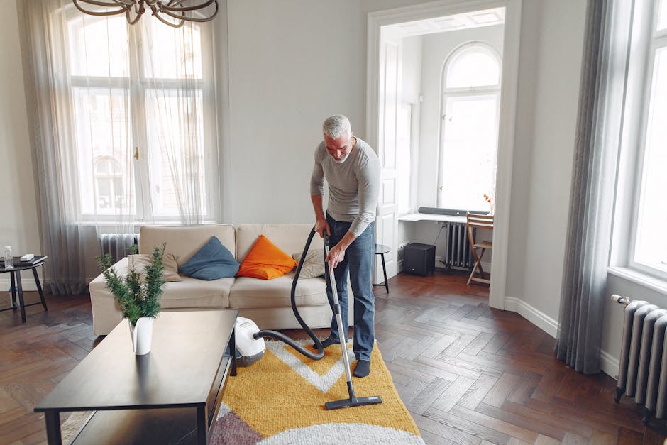 An elderly man with grey hair and glasses is vacuuming a yellow, orange, and white patterned area rug in a bright living room. The room features large windows with sheer white curtains, allowing natural light to illuminate the space. The floor is made of polished dark hardwood, and there is a white sofa with colorful cushions, including orange and blue, positioned against the wall. A small side table holds a glass of water, and underneath a window, a compact black speaker is visible. The walls are painted in a soft light shade, and a white fireplace mantel with a decorative shelf is seen near the windows. The scene reflects surface cleaning and general deep cleaning procedures carried out by [COMPANY_NAME], emphasizing hygiene and maintenance in a domestic setting.