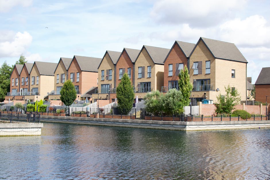 A row of newly built houses with modern brick facades of varying shades, featuring pitched roofs covered with grey tiles. The houses are multi-story, with large windows and small balconies on the upper floors. The front gardens are landscaped with green grass, bushes, and small trees, all enclosed by low brick and metal fences. The properties are situated along a calm waterway with a stone embankment, and the scene is lit by natural daylight under a partly cloudy sky. Cleaners Merton specializes in surface cleaning and deep cleaning for new builds, ensuring premises like these are maintained to high standards of hygiene and appearance.