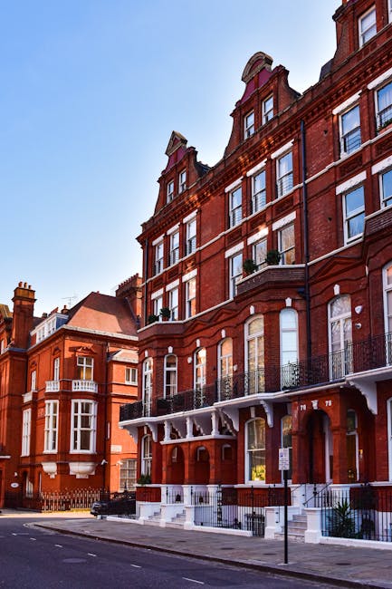A row of newly built houses with modern brick facades of varying shades, featuring pitched roofs covered with grey tiles. The houses are multi-story, with large windows and small balconies on the upper floors. The front gardens are landscaped with green grass, bushes, and small trees, all enclosed by low brick and metal fences. The properties are situated along a calm waterway with a stone embankment, and the scene is lit by natural daylight under a partly cloudy sky. Cleaners Merton specializes in surface cleaning and deep cleaning for new builds, ensuring premises like these are maintained to high standards of hygiene and appearance.