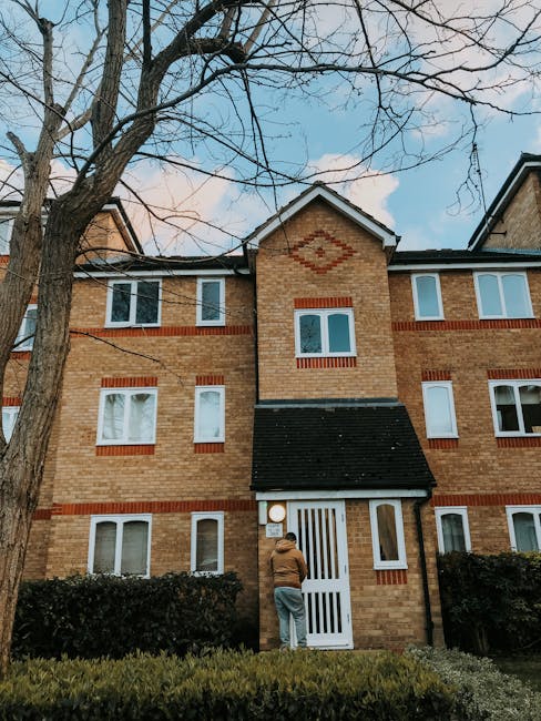 Photograph of a three-story residential building constructed with light brown brickwork accented by red brick details around the windows. The building features multiple white-framed windows, some with visible blinds, and a black-tiled pitched roof. In front, a man with a brown jacket and light-colored pants stands near a white striped gate, which provides access to the entrance. The surrounding area includes well-maintained hedge bushes at the base of the building and a large, leafless tree with sprawling branches against a partly cloudy blue sky. The scene is well-lit, highlighting the clean and tidy appearance of the building facade, indicating recent exterior surface cleaning or maintenance. This setting aligns with the comprehensive domestic and surface cleaning services provided by Cleaners Merton, as outlined in the 'Cleaning Guide for Kingston Road Flats, Raynes Park, Merton'.