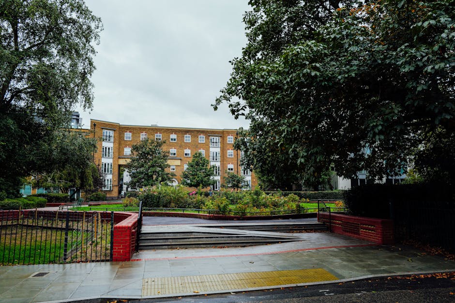 An outdoor scene in a residential area showing a paved pathway leading to a set of steps with black metal railings, surrounded by landscaped garden beds with green shrubs and flowering plants. The pathway is wet, indicating recent cleaning or rain, with a nearby brick wall and a large leafy tree providing shade. In the background, there is a multi-storey brick apartment building with multiple white-framed windows and balconies, under an overcast sky. The scene is well-maintained, demonstrating outdoor surface cleaning and garden care, as provided by Cleaners Merton, aligned with the cleaning guide for Kingston Road flats, Raynes Park, Merton.