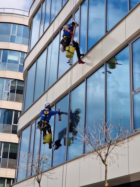 A window cleaner wearing a dark jacket, grey trousers, and a beanie is seated on a safety harness while cleaning large glass windows of a modern commercial building. The worker is using a squeegee to 