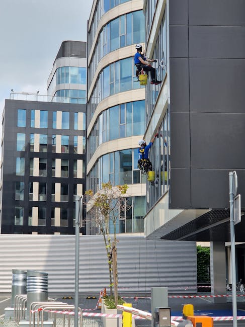 Two window cleaners wearing safety helmets and harnesses are performing surface cleaning on the exterior glass windows of a modern multi-story office building. They are using squeegees and cloths, wit