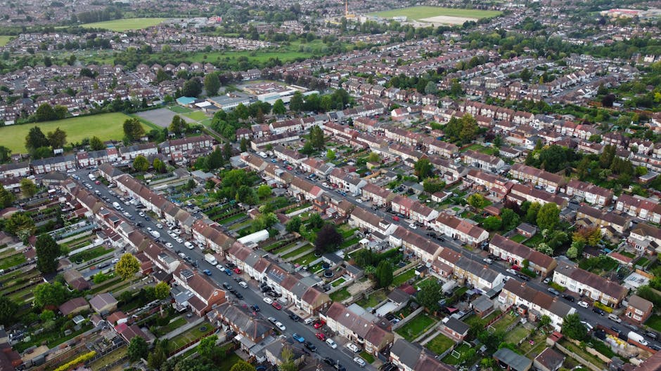 Aerial view of a suburban residential neighborhood in Colliers Wood, Merton, showing rows of terraced houses with tiled roofs, small front gardens, and parked cars lining the streets. The area includes tree-lined streets with lush greenery interspersed between homes, and open green spaces and sports fields visible in the background under a slightly overcast sky, captured during daytime. This image reflects the peaceful, well-maintained environment where professional domestic cleaning services like those offered by Cleaners Merton can help maintain hygiene and tidiness.