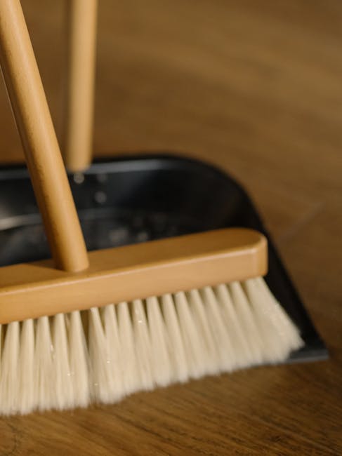 Close-up image of a cleaning broom with a wooden handle and a beige plastic head, fitted with synthetic bristles, resting on a wooden floor. The broom appears clean and well-maintained, and is positioned near a dark-colored dustpan, which is partially visible in the background. The scene is illuminated with natural or ambient light, highlighting the smooth surface of the wooden flooring. This image exemplifies surface cleaning tools used in domestic or commercial settings, aligning with the services offered by Cleaners Merton for end-of-tenancy and deep cleaning in Colliers Wood, Merton, to ensure hygiene and spotless surfaces.