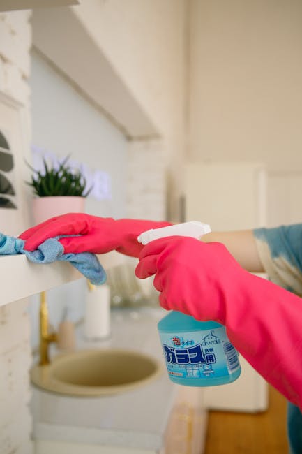 Close-up of a person wearing pink rubber gloves and using a spray bottle to sanitise a white kitchen shelf, which holds a small potted plant with green foliage. The shelf appears to be made of glossy white material, and the background includes a cream-colored brick wall and a sink with a beige drain. The scene demonstrates surface cleaning and sanitisation processes in a domestic kitchen, highlighting hygiene and cleanliness standards often emphasized by Cleaners Merton. Natural lighting enhances the bright, orderly appearance of the space, reflecting a well-maintained home environment suitable for deep cleaning services.