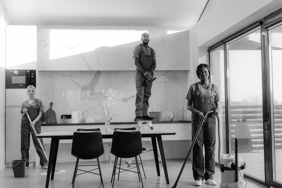 A black-and-white photograph of three professional cleaners in a modern kitchen, part of a domestic cleaning service by Cleaners Merton. The kitchen features a large, polished white marble backsplash with grey veining, a smooth countertop, and large sliding glass doors allowing natural light to fill the space. One cleaner is standing on a step ladder, wiping down the upper cabinets; another cleaner is holding a mop near the floor, which appears spotless; the third is holding a spray cloth, indicating surface cleaning; the room is tidy with minimal clutter, and the lighting highlights the pristine, shiny surfaces, emphasizing the company's focus on deep cleaning and hygiene maintenance in residential environments.