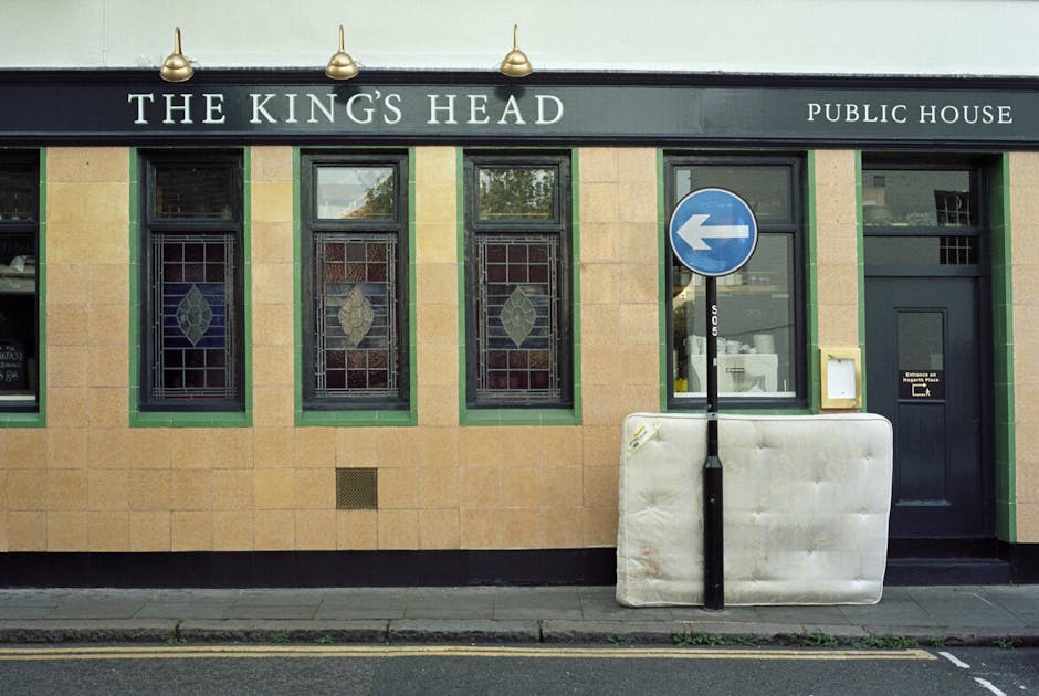 An overflowing public recycling and waste collection point in an urban area, featuring a large grey metal bin labeled for paper and card waste, with its lid open and surrounded by discarded cardboard 