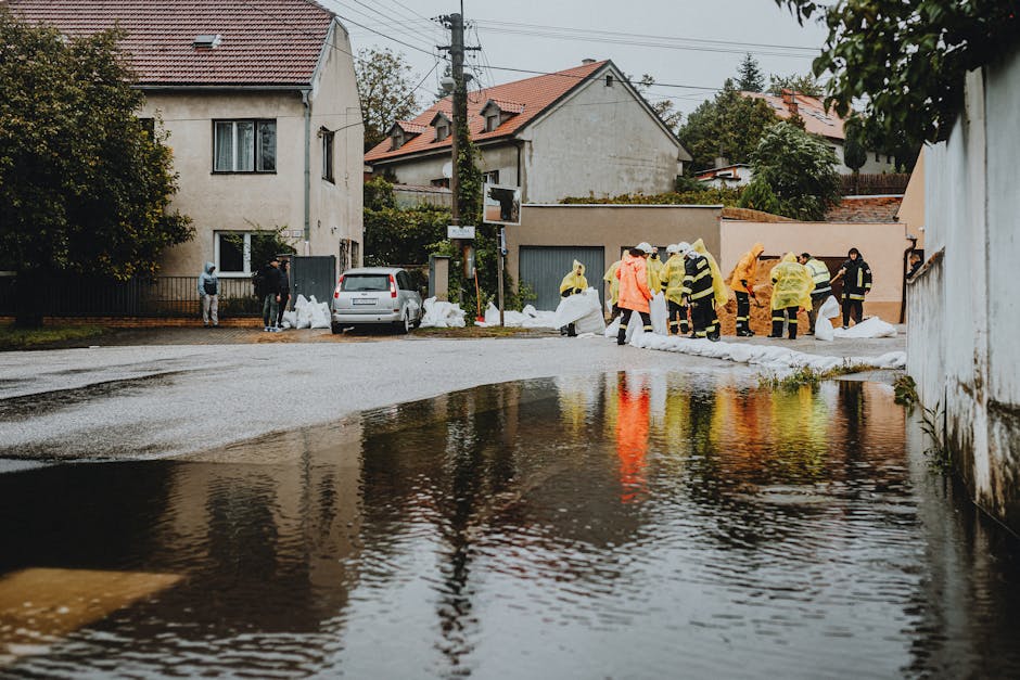 A group of emergency responders in bright yellow and orange waterproof jackets and reflective gear are working outdoors in a residential area during a flood cleanup. They are gathered along the edge of a wet street, which is partially submerged with water pooling on the surface. Some responders are handling sandbags to prevent further water intrusion, while others are using tools to remove debris from the area. In the background, there are houses with tiled roofs, a parked car, and trees. The scene is overcast, with soft natural lighting highlighting the damp surfaces and the effort to restore safety and cleanliness, demonstrating surface cleaning and flood mitigation efforts by Cleaners Merton in response to urgent flood situations.