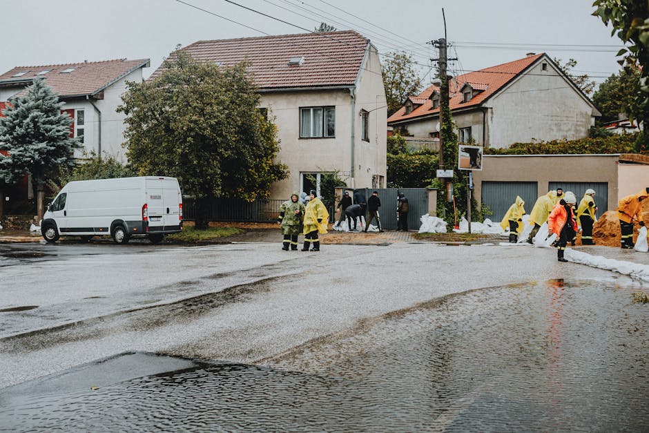 A street scene viewed outdoors with wet, reflective asphalt demonstrating recent flooding. Several workers wearing yellow and orange waterproof suits and protective gear are engaged in flood cleanup efforts, including placing sandbags along the curb. A white utility van is parked on the roadside, while residential houses with tiled roofs and trees line the background. The lighting suggests overcast weather, contributing to the damp, pristine condition of the surfaces. The image captures the collaborative process of deep cleaning and flood mitigation, highlighting the importance of sanitation and surface cleaning in such emergency responses. The scene is associated with Cleaners Merton's urgent flood cleanup services in Merton, emphasizing professional intervention in maintaining hygiene and safety after flooding incidents.