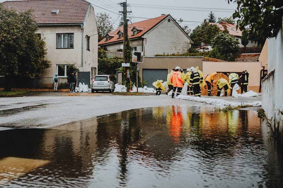 A group of emergency responders in bright yellow and orange waterproof jackets and reflective gear are working outdoors in a residential area during a flood cleanup. They are gathered along the edge of a wet street, which is partially submerged with water pooling on the surface. Some responders are handling sandbags to prevent further water intrusion, while others are using tools to remove debris from the area. In the background, there are houses with tiled roofs, a parked car, and trees. The scene is overcast, with soft natural lighting highlighting the damp surfaces and the effort to restore safety and cleanliness, demonstrating surface cleaning and flood mitigation efforts by Cleaners Merton in response to urgent flood situations.